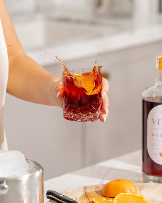 Person holding a glass of red cocktail with orange slices and a bottle in the background